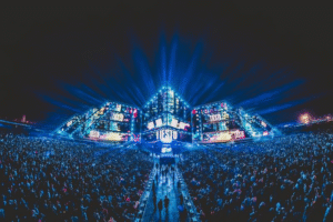 Tiesto performing at a large outdoor night music festival with vibrant lighting and a massive crowd