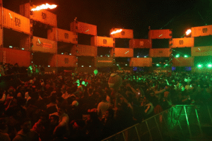 A vibrant nighttime music festival with a large crowd and dynamic stage effects featuring flames and container structures